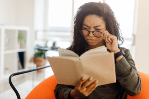 When Your Glasses Prescription Is Not Working: How to Fix It, Focused confused young black lady trying to read paper book, squinting to see more clearly, wearing glasses, having difficulties seeing text because of vision problems, sitting on pouf in living room
