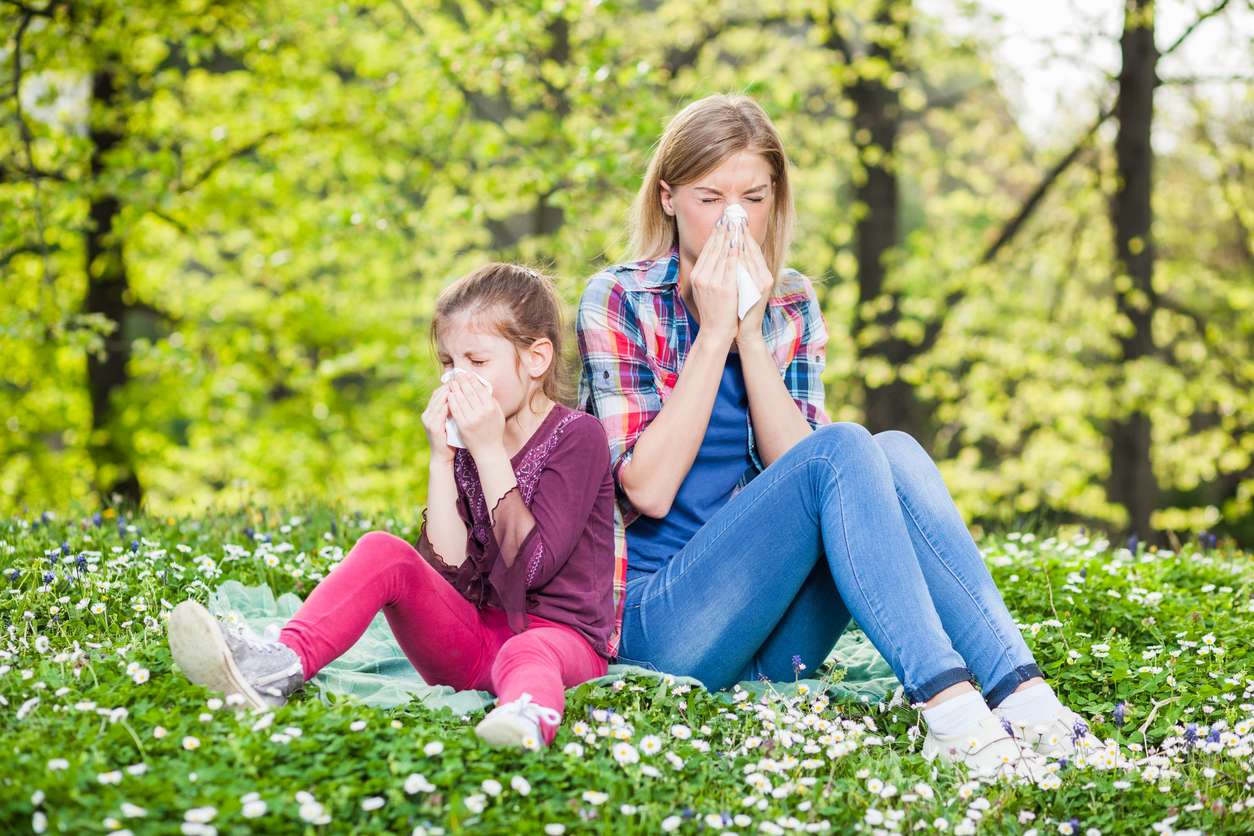 Spring Allergies & Your Eyes: A Texas Family Eye Care Guide, Woman and child blowing their noses into tissues while sitting in a field of flowers, indicating spring allergies or hay fever.