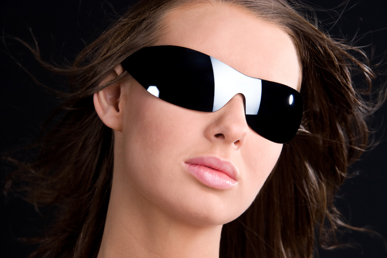 Family Eye Care, Close-up portrait of a woman with windswept brown hair wearing black futuristic shield sunglasses and glossy lips.