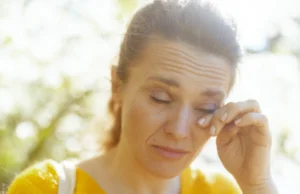 Woman rubbing her eye outdoors in bright summer sunlight, illustrating dry, irritated eyes and summer eye discomfort.